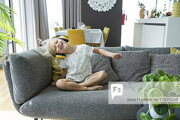 Smiling girl listening music through wireless headphones sitting on sofa in living room