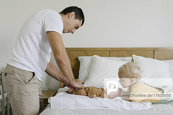 Boy looking at father buttoning baby's rompers on bed
