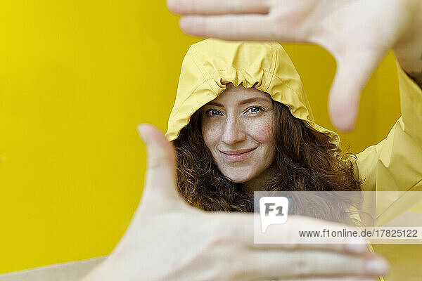Smiling woman in yellow raincoat making frame with hands in front of wall