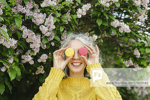 Happy woman covering face with macaroons in front of lilac at park
