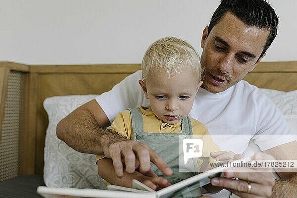 Father reading book with son sitting on bed at home