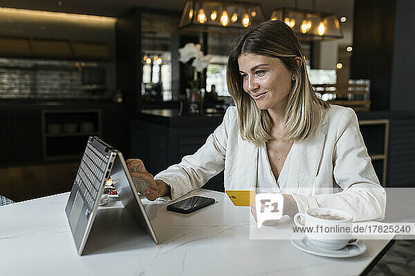 Smiling businesswoman holding credit card doing online shopping through laptop at restaurant
