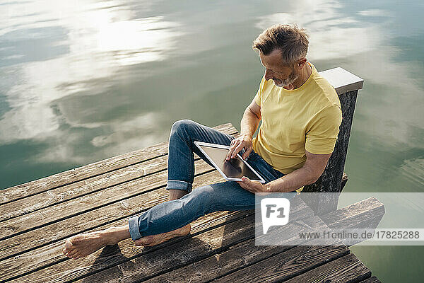 Mature man using tablet PC sitting on pier over lake