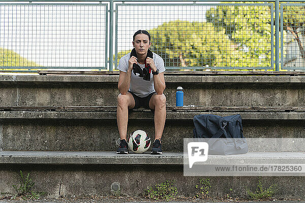 Selbstbewusste junge Frau mit Fußball sitzt auf der Bank am Sportplatz