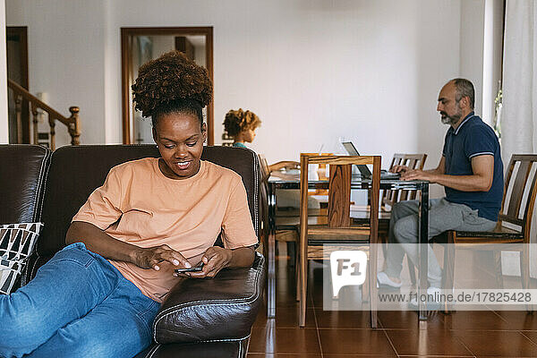 Lächelnde Frau sitzt mit Smartphone vor Mann und Tochter zu Hause