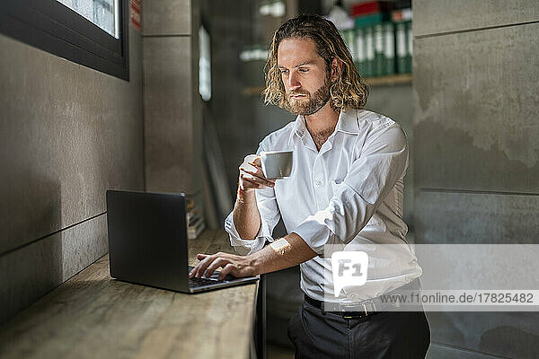 Serious businessman having coffee using laptop in office