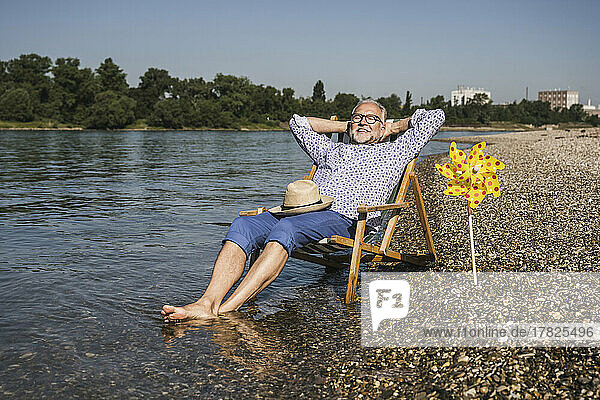 Smiling man with hands behind head sitting by paper pinwheel toy at riverbank