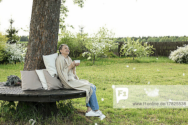 Smiling woman wrapped in blanket holding cup sitting on bench around tree trunk