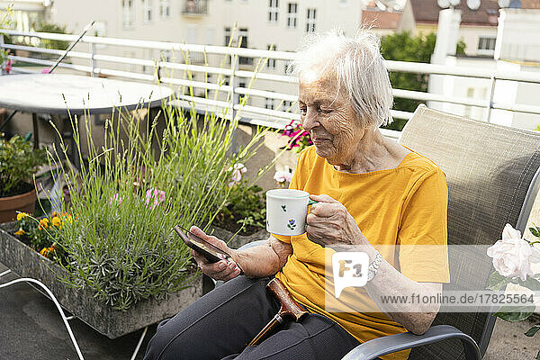 Smiling senior woman holding coffee cup looking at mobile phone on balcony