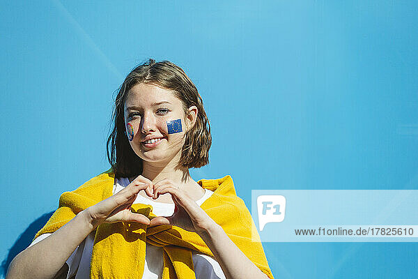 Smiling teenage girl with peace symbol and European Union paint on cheeks gesturing heart shape in front of blue wall