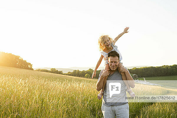 Smiling man carrying daughter sitting with arms outstretched at field