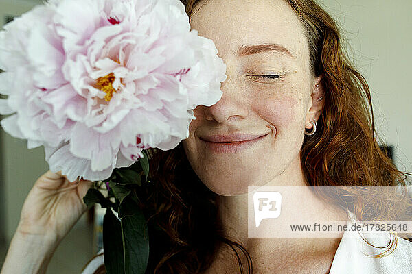 Woman with eyes closed holding pink flower in front of eye