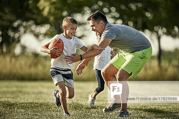 Cheerful man chasing son running with rugby ball at sports field