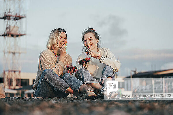 Happy mother and daughter eating strawberries sitting at beach