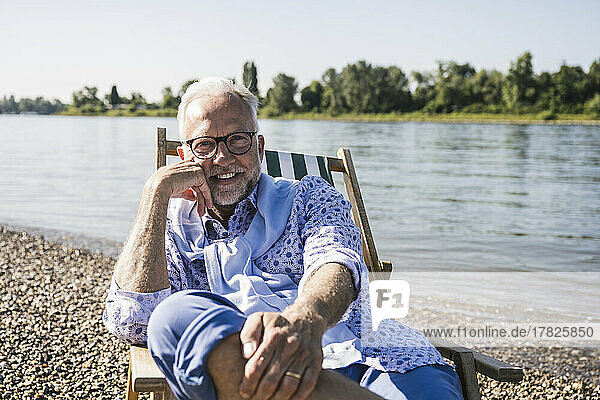 Smiling man wearing eyeglasses sitting on deck chair at riverbank