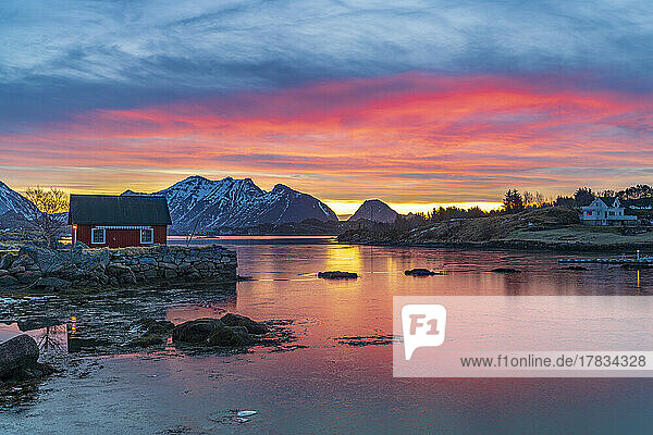 Feuriger Himmel bei Sonnenaufgang über einem roten Rorbu mit Blick auf das gefrorene Meer  Ballstad  Vestvagoy  Lofoten  Norwegen  Skandinavien  Europa