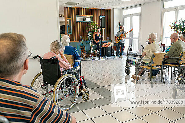 Seniors in a retirement home sitting on chairs or wheelchairs to listen to a concert.