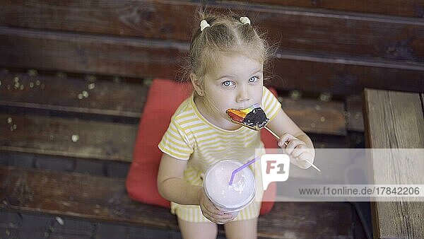 Ittle Mädchen isst einen bunten Lebkuchen und hält einen Milchshake in der Hand. Nahaufnahme eines süßen Mädchens  das auf einer Parkbank sitzt und Kekse mit einem Milchshake isst und in die Linse der Kamera schaut. Ansicht von oben. Odessa  Ukraine  Europa