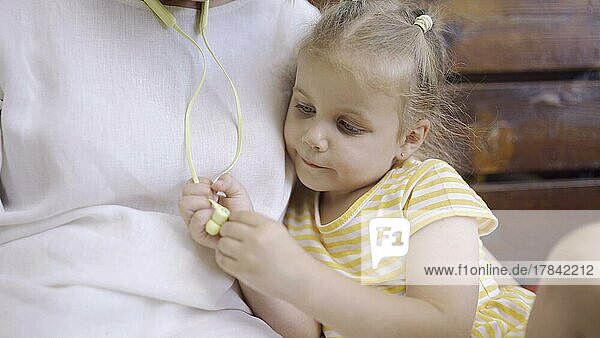 Nahaufnahme von niedlichen kleinen Mädchen sitzt neben Mama und spielt mit ihren Kopfhörern. Nahaufnahme von Kind Mädchen sitzt auf Parkbank und untersucht Mütter Kopfhörer. Odessa  Ukraine  Europa