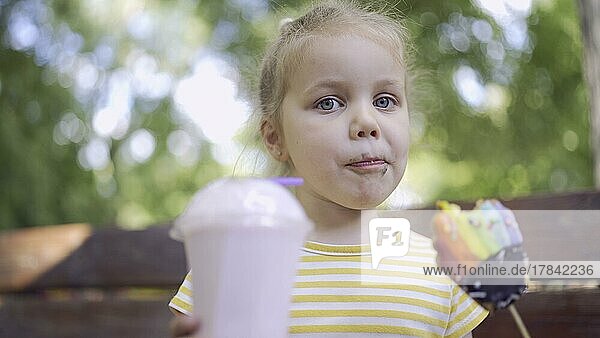 Ittle Mädchen isst einen bunten Lebkuchen und hält einen Milchshake in der Hand. Nahaufnahme eines süßen Mädchens  das auf einer Parkbank sitzt und Kekse mit einem Milchshake isst und in die Linse der Kamera schaut. Odessa  Ukraine  Europa