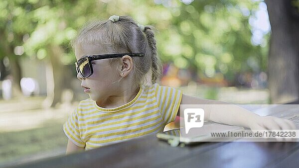 Niedliches kleines Mädchen mit Sonnenbrille hört Musik auf einem Mobiltelefon und singt mit. Close-up-Porträt von Kind Mädchen sitzt in Sonnenbrille auf Stadtpark und hört Musik mit Handy. Odessa  Ukraine  Europa