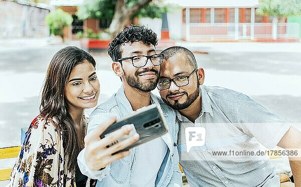Drei lächelnde Freunde sitzen auf einer Bank und machen ein Selfie. Vorderansicht von drei glücklichen Freunden  die auf einer Bank sitzend ein Selfie machen. Drei Freunde machen ein Selfie auf einer Parkbank