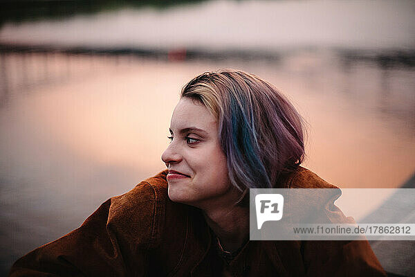 Portrait of happy young woman sitting by river in city at sunset