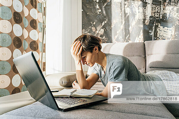 Woman annoyedly holds on to her head lying on the couch near laptop
