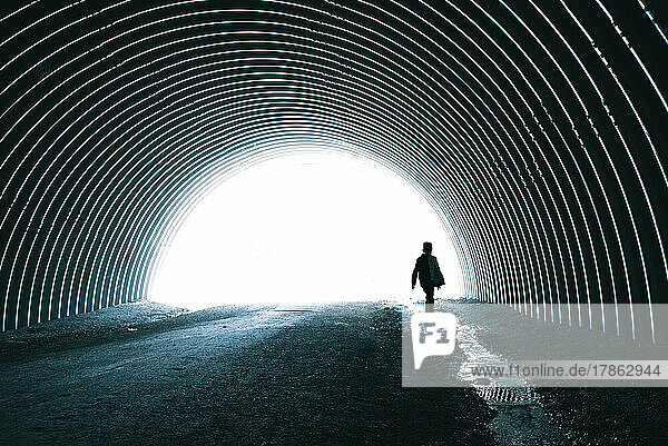 Child walking alone beside stream running through dark metal tunnel.