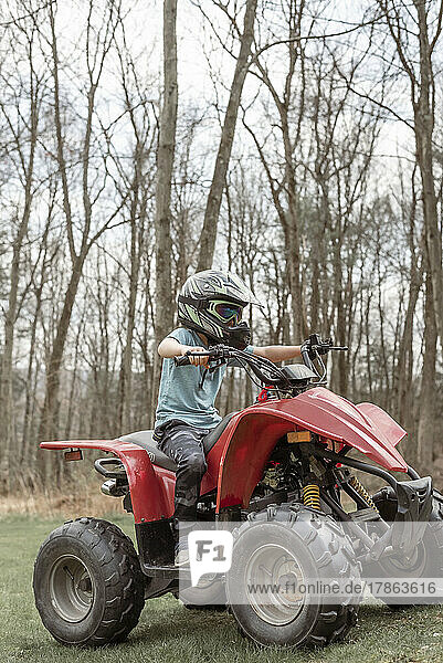 Boy riding a red four wheeler in grass with woods in background