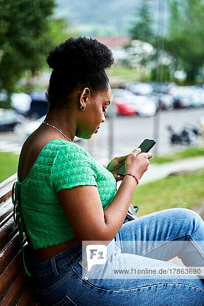 Afro american woman sitting on bench with mobile phone in park