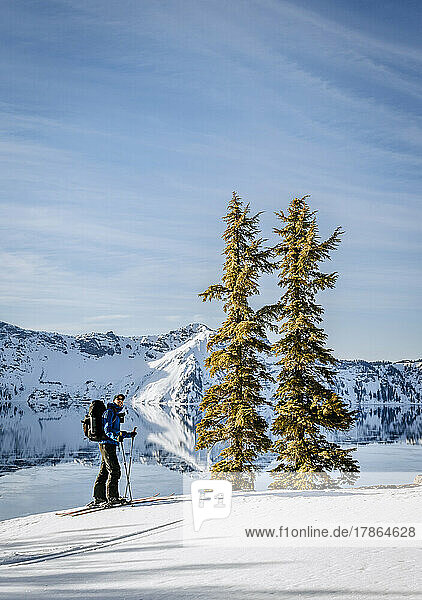 Skier takes in view of Crater Lake and two pine trees in winter.