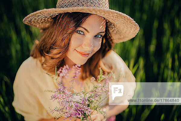 Pretty countryside woman in straw hat with flowers bouquet in field.