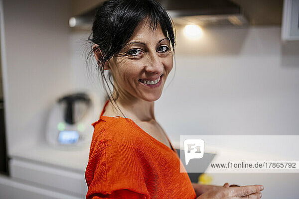 Smiling woman in orange sweater standing at kitchen   looking camera