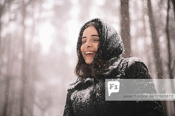 Portrait of happy young woman standing in forest during snowfall