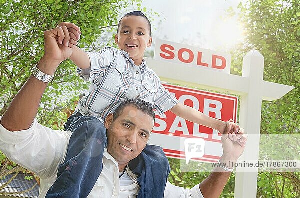 Glücklicher junger hispanischer Vater und Sohn vor einem zum Verkauf stehenden Immobilienschild