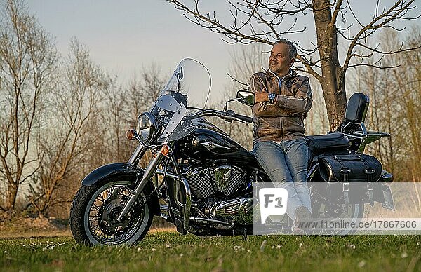 Glücklicher Mann im mittleren Alter  lehnt sich fröhlich und zufrieden an sein Motorrad  Karlsruhe  Deutschland  Europa