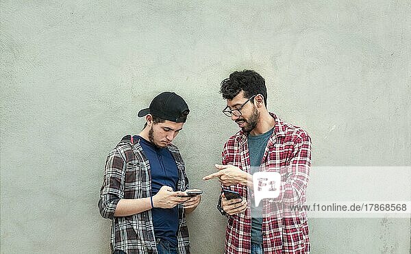 Zwei junge Freunde  die an einer Wand lehnen und ihre Handys überprüfen  Zwei Freunde  die an einer Wand lehnen und auf den Inhalt ihrer Handys schauen. Freund zeigt seinem Freund im Freien sein Handy