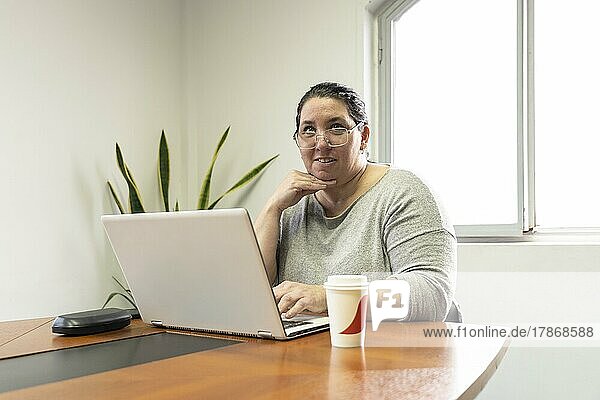 Reife Geschäftsfrau sitzt am Kopfende eines Tisches in einem Besprechungsraum  arbeitet an ihrem Computer und sieht jemanden an  der außerhalb des Bildes steht