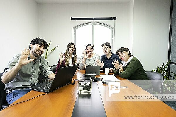 Gruppe von Mitarbeitern sitzen im Büro Besprechungsraum Blick in die Kamera und winken. Online Meeting Konzept