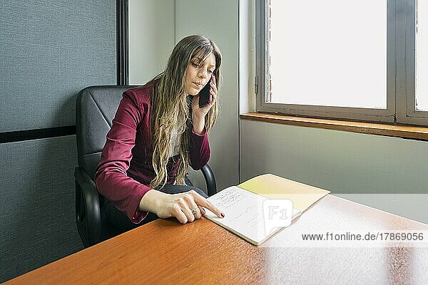 Junge blonde Geschäftsfrau sitzt in ihrem Büro und telefoniert  während sie Notizen überprüft