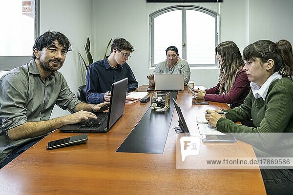 Eine Gruppe von Angestellten sitzt um den Besprechungstisch im Büro und arbeitet an ihren Notebooks  einer von ihnen schaut in die Kamera