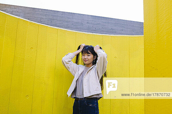 Lächelnde junge Frau  die vor gelber Wand Haare bindet