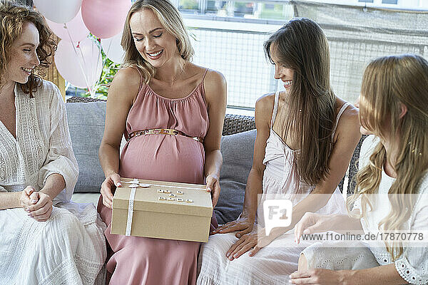 Smiling pregnant woman opening gift box by friends on sofa