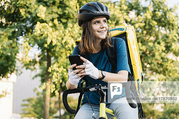 Smiling delivery woman wearing helmet sitting with smart phone on bicycle