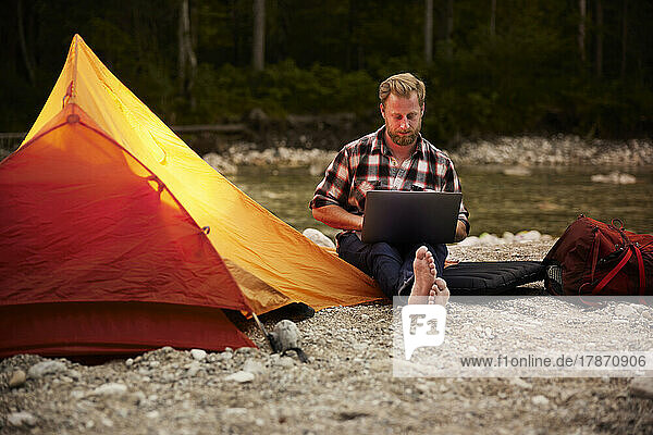 Businessman using laptop sitting by tent at riverbank