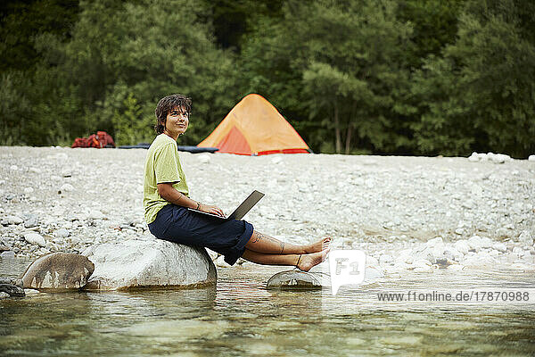 Smiling businesswoman with laptop sitting on rock at riverbank