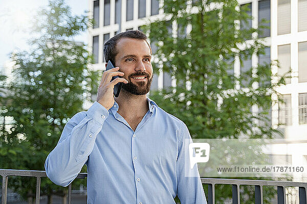 Happy businessman talking on smart phone standing in front of trees