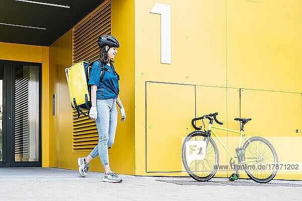 Smiling young delivery person with backpack walking towards bicycle