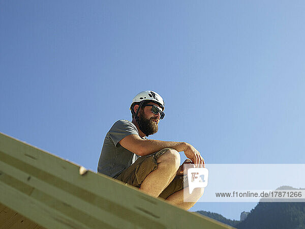 Carpenter wearing hardhat sitting on roof under blue sky